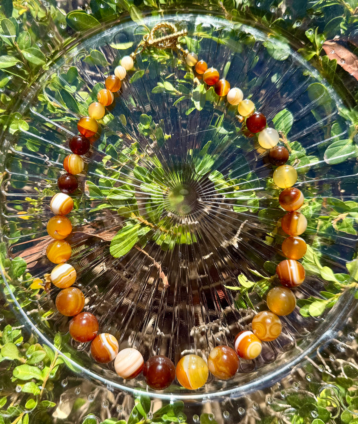 Top-down view of a natural banded agate gradient necklace on a clear vintage glass plate, showing earth-toned round beads graduating in size with a gold toggle clasp.
